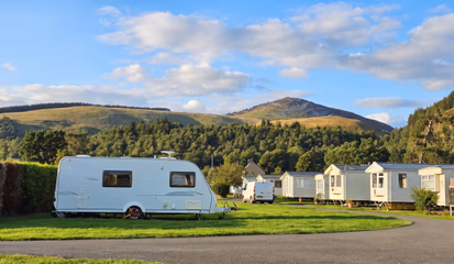 A touring caravan pitched on grass beside a hedge, with static caravans and a hillside beyond