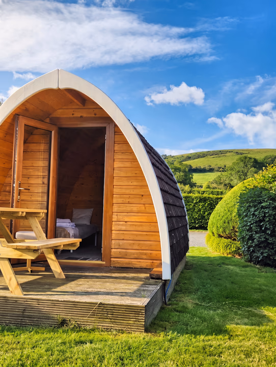 A timber camping pod with its door open and a picnic bench on the decking, looking out over open hillside