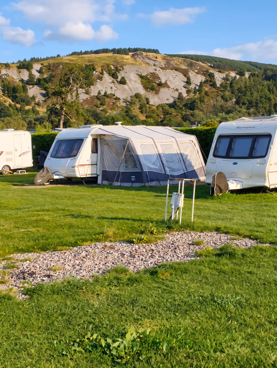 A touring caravan with its awning up on a long-stay pitch below a wooded hillside