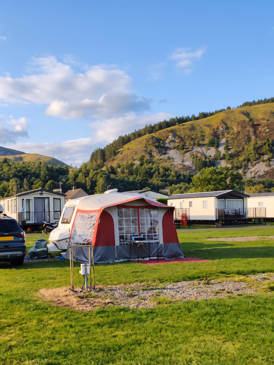 A touring caravan with a red and grey awning on a grass pitch, static caravans and hills behind